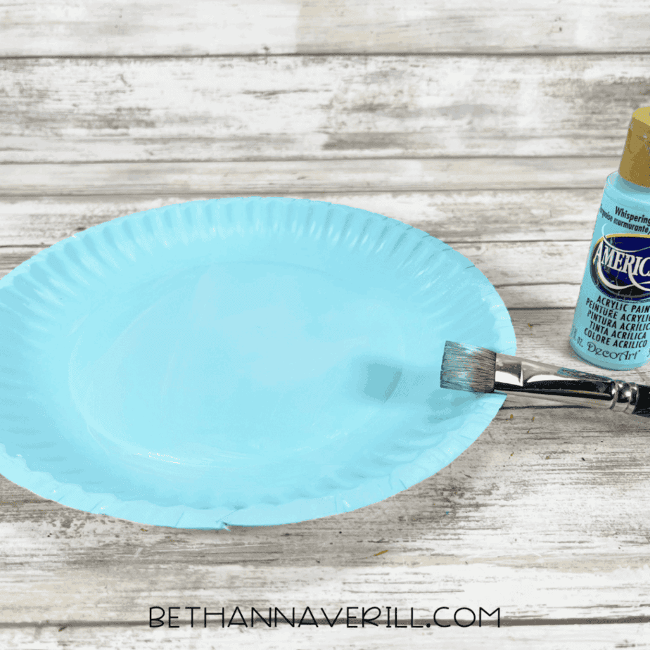 Paper plate painted light blue with acrylic paint and a paintbrush beside it.