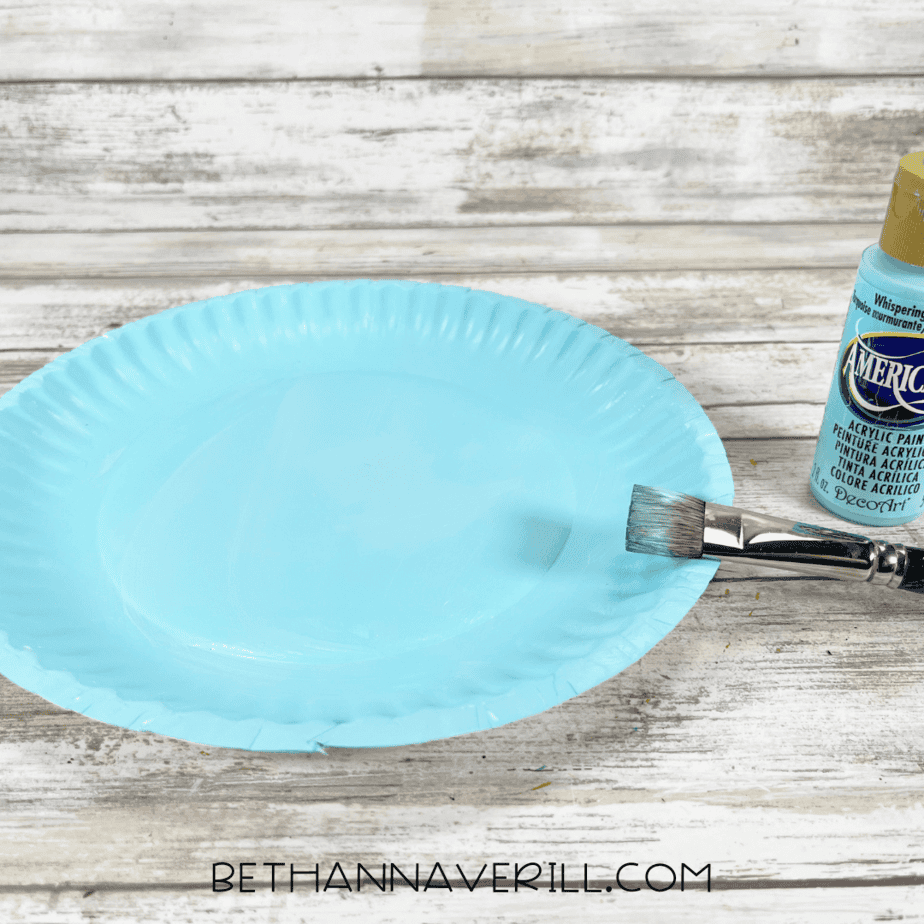 Paper plate painted light blue with acrylic paint and a paintbrush beside it.