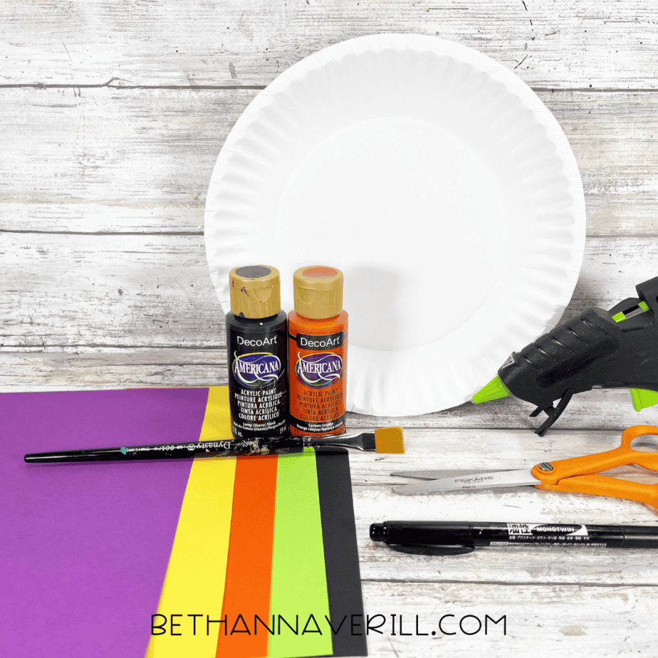 Materials for witch hat paper plate craft including paint, paper plate, glue gun, and construction paper.