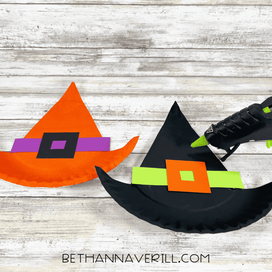 Two witch hats, one black and one orange, with colored hat bands and buckles being glued on.