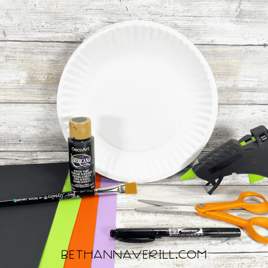 Materials for witch legs paper plate craft including paint, glue gun, scissors, and colorful paper.