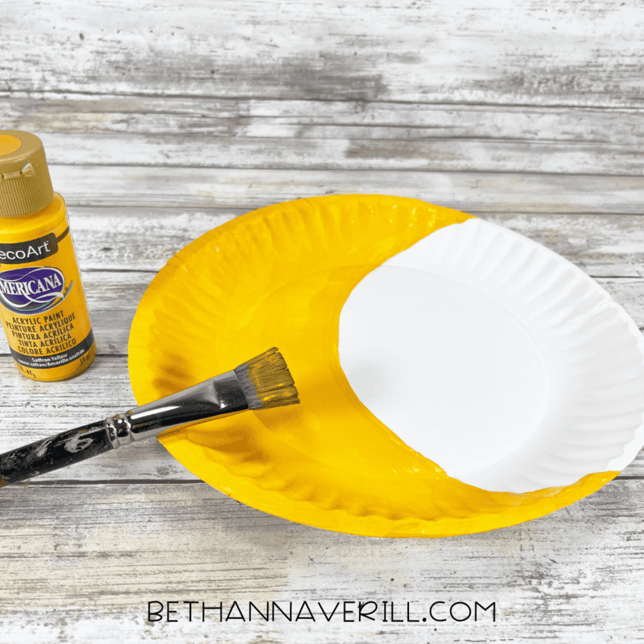 Paper plate half painted yellow for the moon with paintbrush and paint nearby.