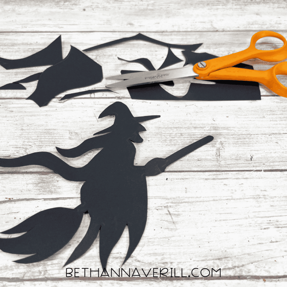 Cut-out black paper witch shape beside scissors and scrap paper pieces.