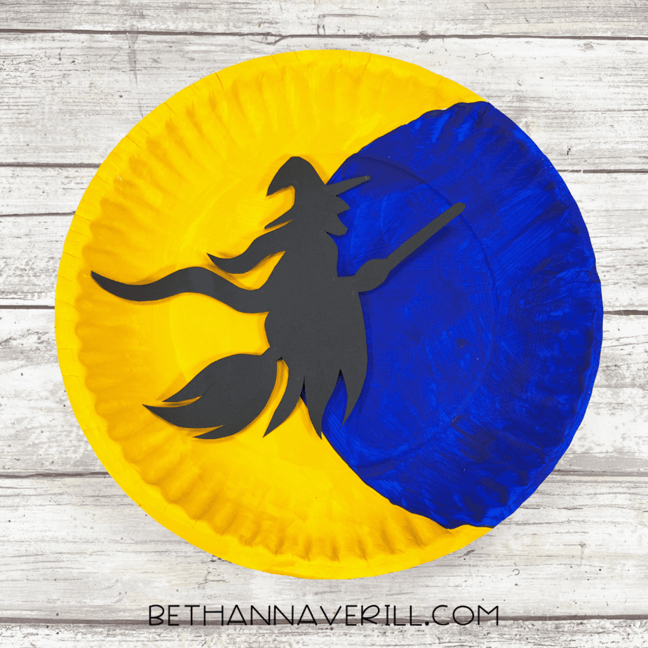 Paper plate painted yellow and blue with witch silhouette placed across moon.