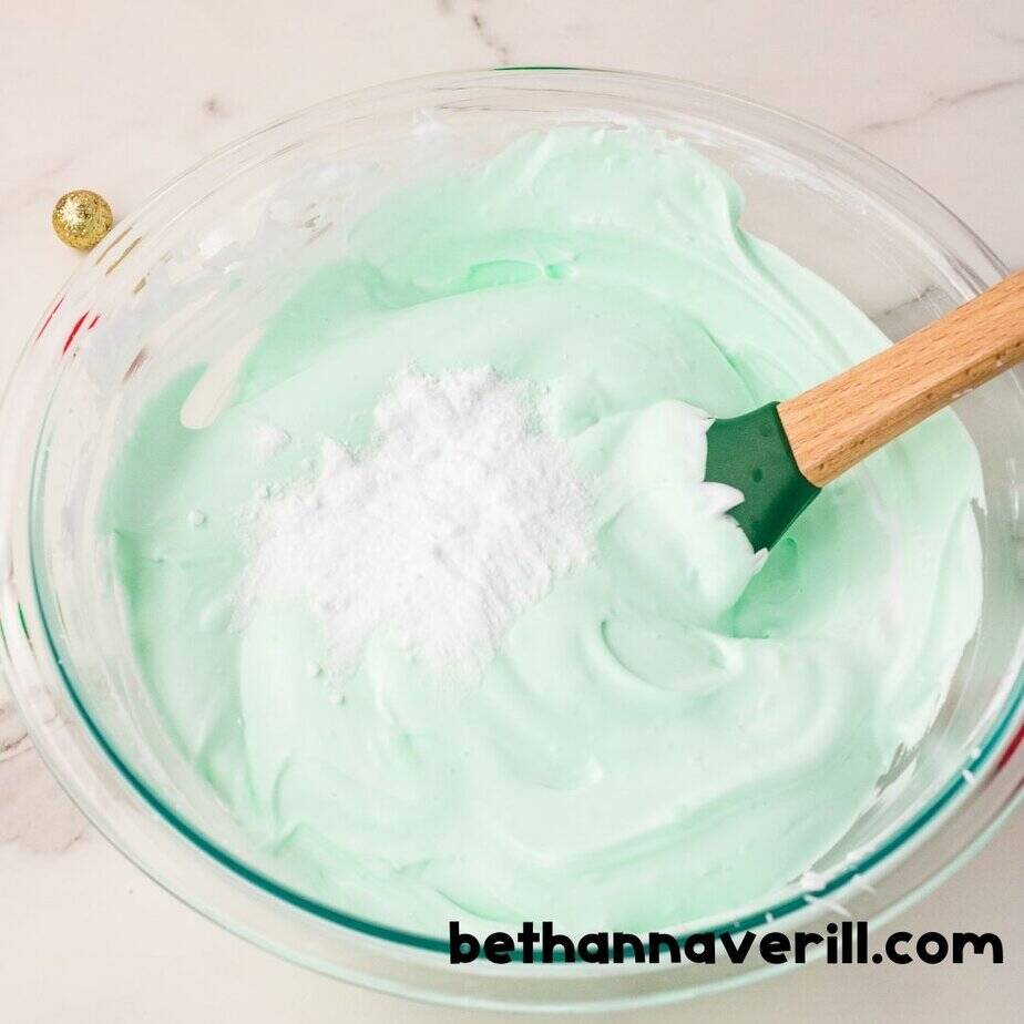 A mound of white baking soda sits on top of whipped green slime in a glass bowl, just before being stirred into the Grinch slime mixture.