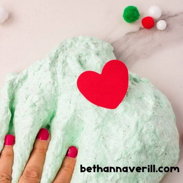 A hand with pink-painted nails presses into fluffy, pale green Grinch slime with red glitter and a red foam heart on top. Red, green, and white pom-poms are scattered in the background.