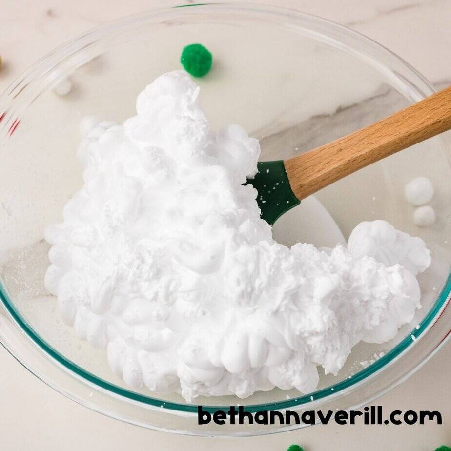 A large pile of white shaving cream sits atop glue in a glass bowl with a green spatula, ready to be mixed to create fluffy Grinch slime.