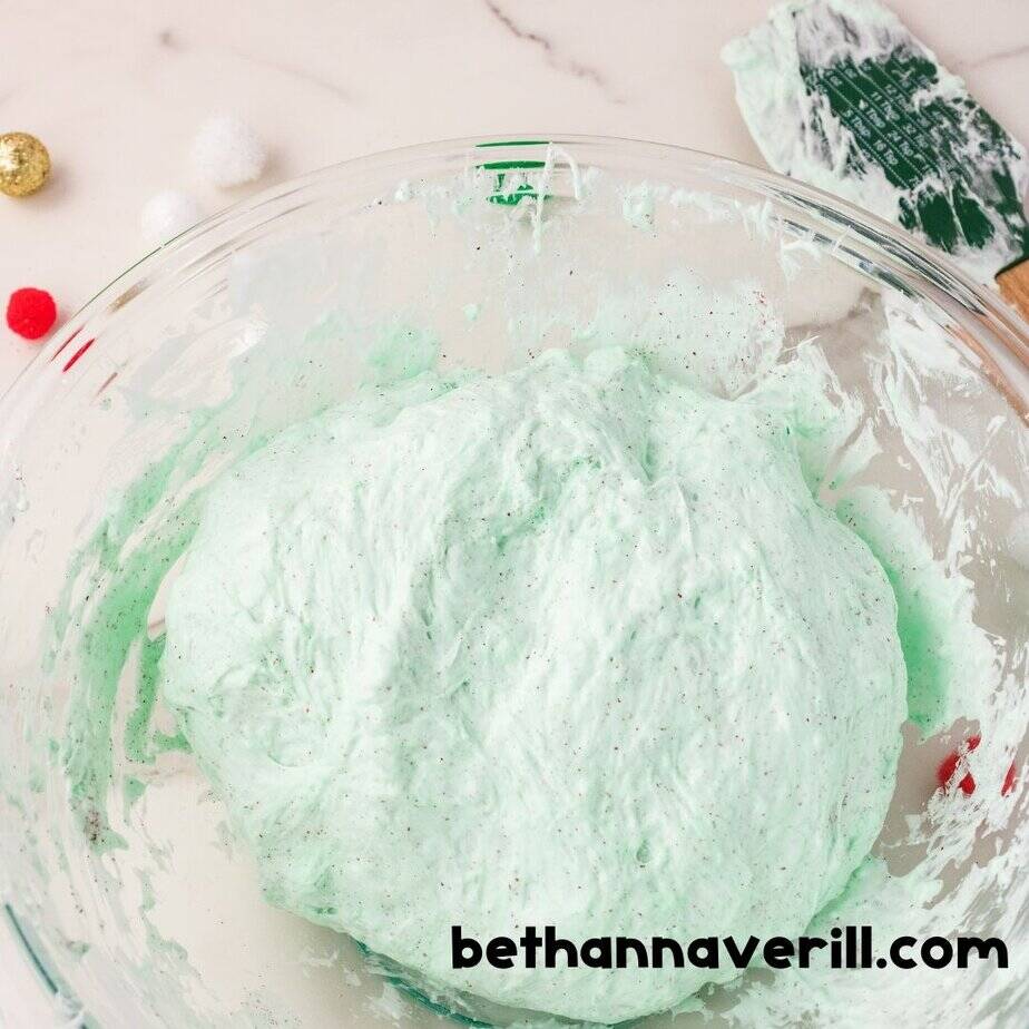 Fully mixed and textured green Grinch slime in a glass bowl with a used spatula nearby. Red and white pom-poms are scattered around the bowl.