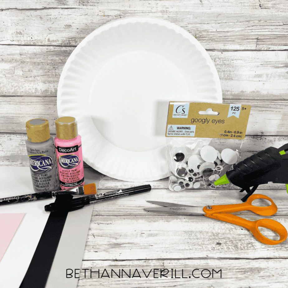 A flat lay of craft materials including a white paper plate, gray and pink acrylic paint, a pack of googly eyes, black and white cardstock, scissors, glue gun, paintbrush, and a black marker on a wooden background.