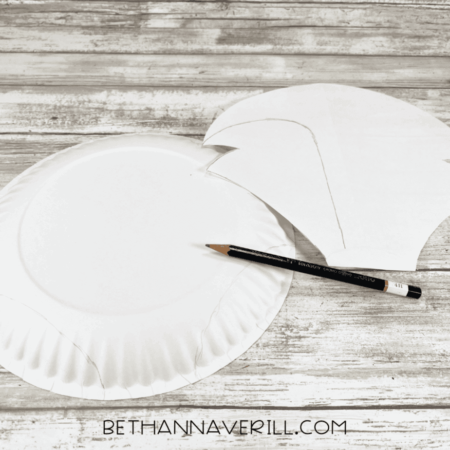 A white paper plate with pencil-traced outlines of the Arctic fox shape, next to a paper pattern and a pencil on a wooden surface.