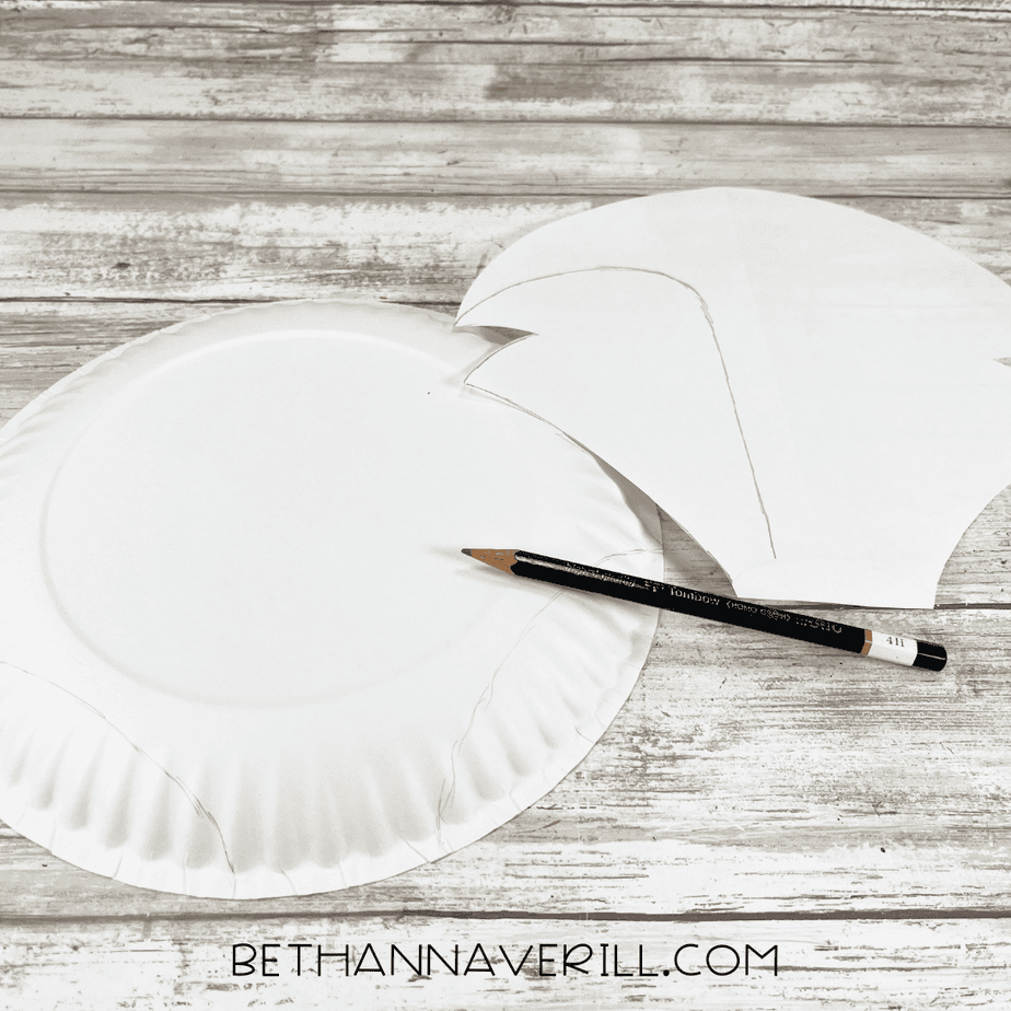 A white paper plate with pencil-traced outlines of the Arctic fox shape, next to a paper pattern and a pencil on a wooden surface.