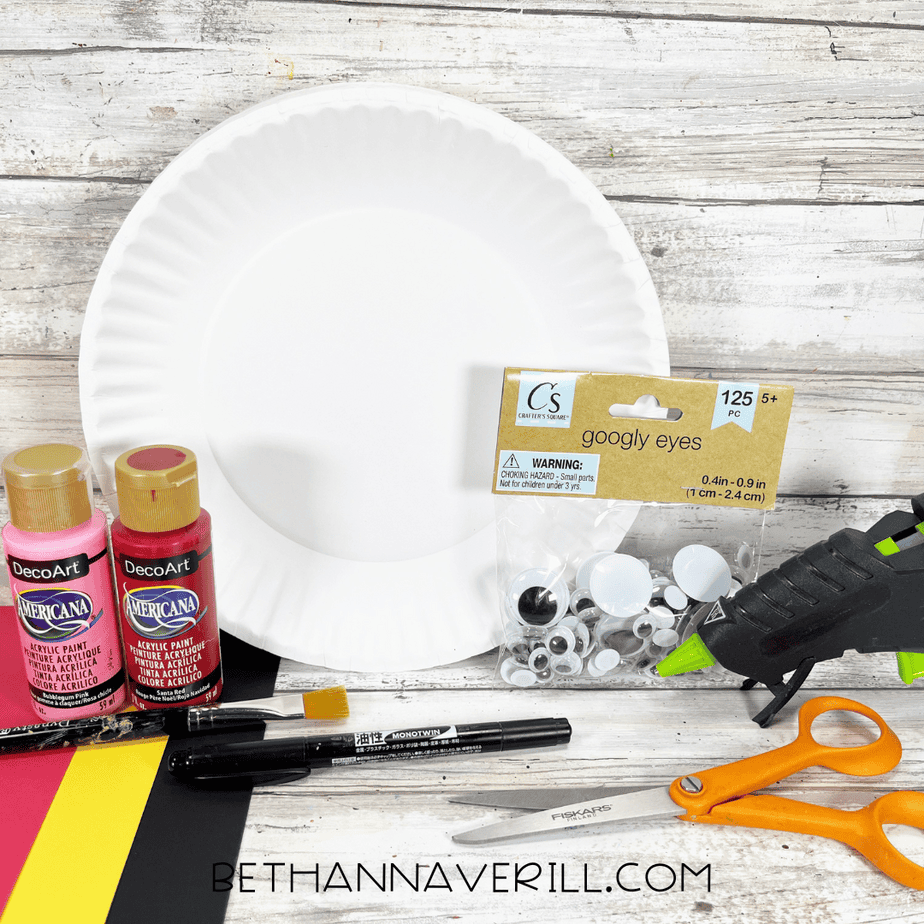 A set of craft supplies for a cardinal paper plate craft, including a blank paper plate, red and pink acrylic paint, googly eyes, scissors, glue gun, paintbrush, and sheets of colored cardstock on a wooden background.