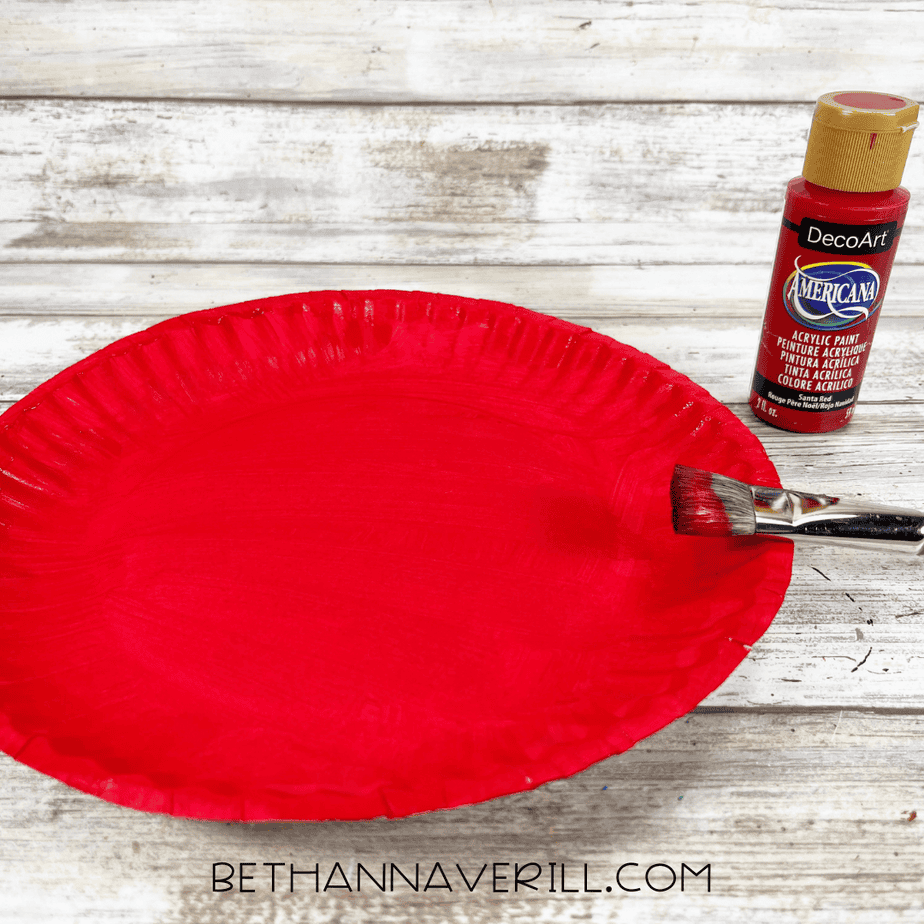 A paper plate painted bright red with a paintbrush resting on the edge and a bottle of red acrylic paint nearby.