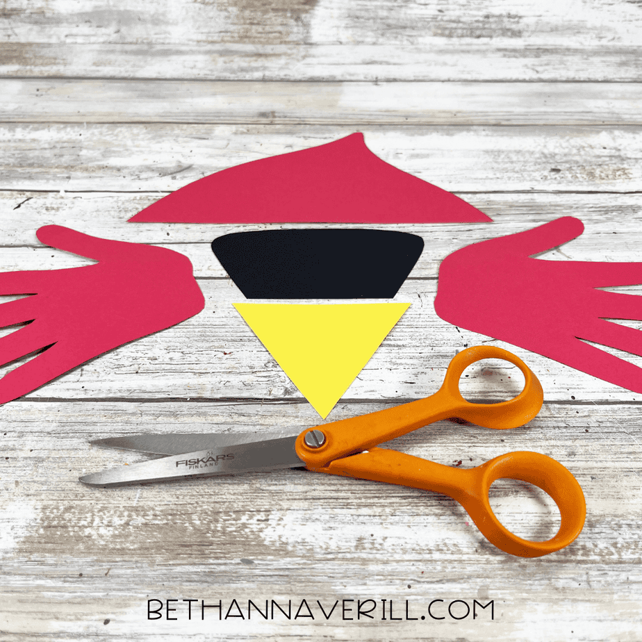 Cut-out cardinal craft pieces laid out on the table, including two red handprint wings, a black face piece, and a yellow triangle beak.