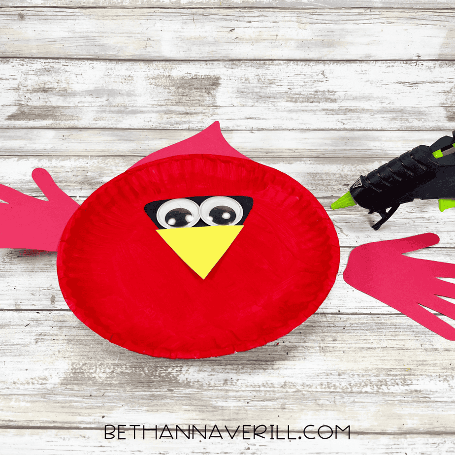 A red paper plate with the black face shape, googly eyes, and yellow beak glued onto the center, with a glue gun beside it.