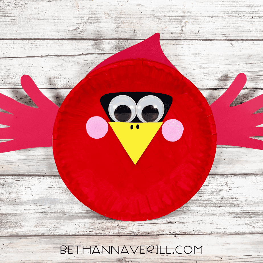 The completed cardinal paper plate craft with googly eyes, yellow beak, pink cheeks, red crest, and handprint wings attached on both sides.