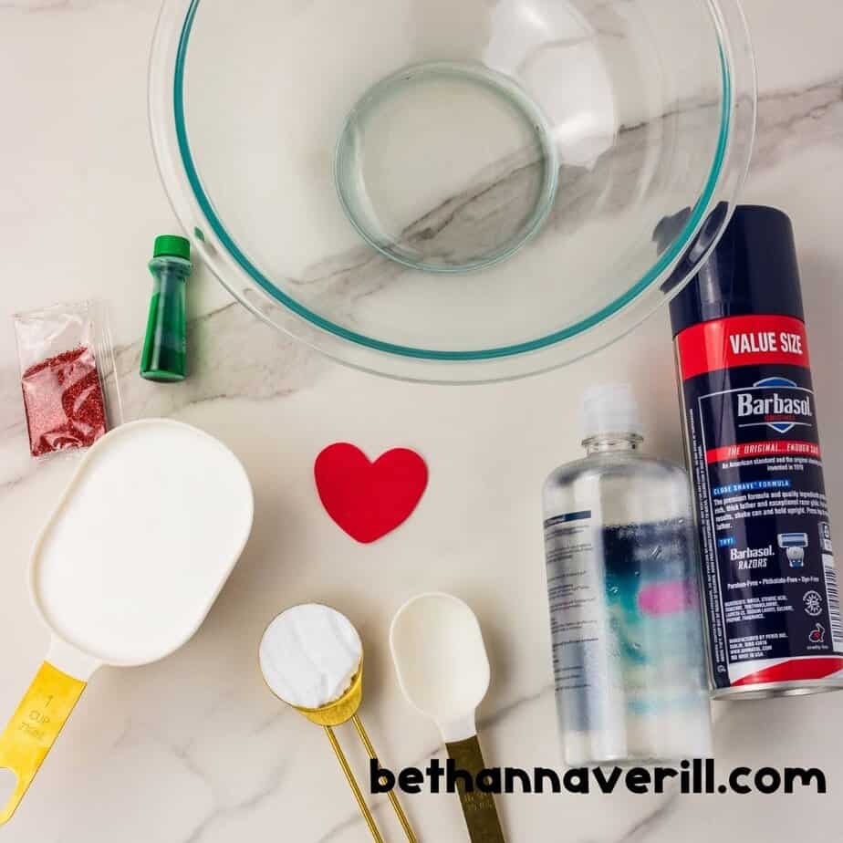 Top-down view of Grinch slime ingredients including glue, baking soda, saline solution, shaving cream, green food coloring, red glitter, and a red heart next to a mixing bowl.