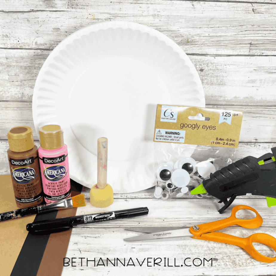 Flat lay of moose craft supplies including a paper plate, brown and pink paint, sponge dabber, googly eyes, glue gun, scissors, marker, and brown cardstock.