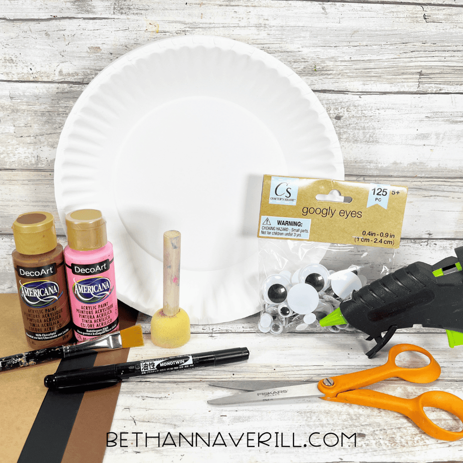 Flat lay of moose craft supplies including a paper plate, brown and pink paint, sponge dabber, googly eyes, glue gun, scissors, marker, and brown cardstock.