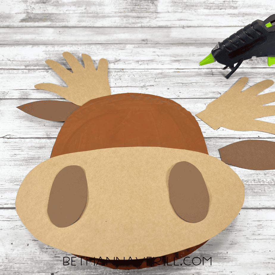 Paper plate moose head with muzzle and ear pieces glued on, and the glue gun visible nearby.