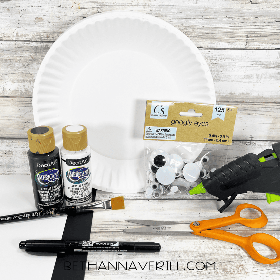 Supplies laid out for the orca paper plate craft, including a white paper plate, black and white paint, googly eyes, black and white cardstock, glue gun, scissors, pencil, and paintbrush.