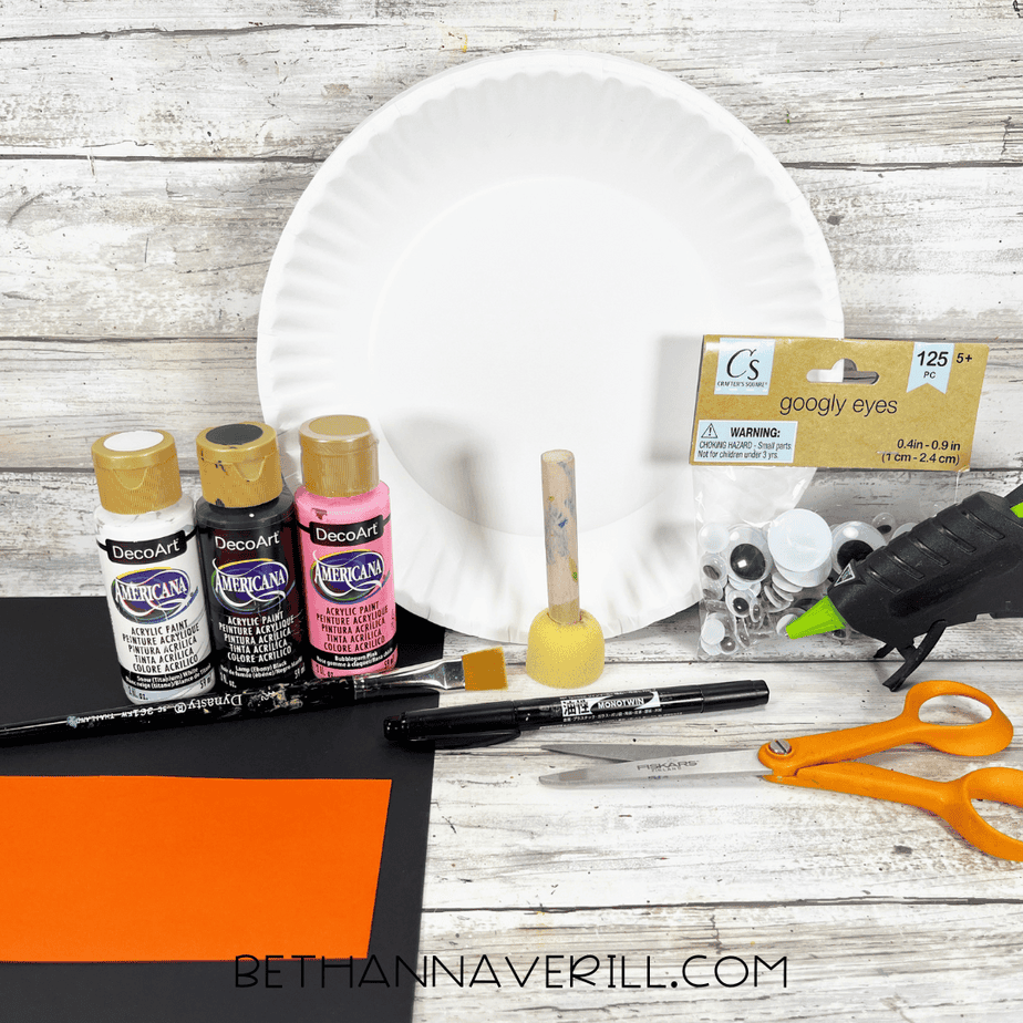 Craft supplies for a paper plate penguin craft including a paper plate, acrylic paints, googly eyes, glue gun, scissors, black and orange cardstock, sponge dabber, paintbrush, and a black marker arranged on a wooden background.