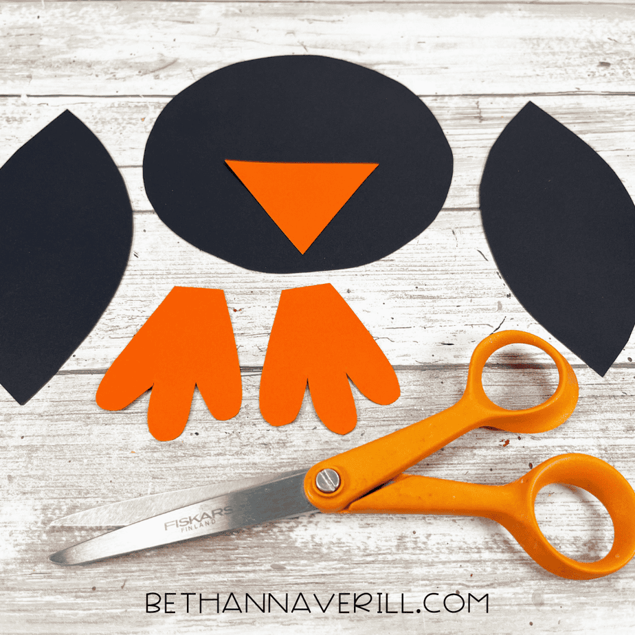 Cut-out penguin pieces including the black head, wings, orange beak, and orange feet arranged next to scissors on a wooden background.