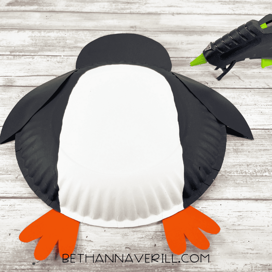 Paper plate with black wings and orange feet glued on, beginning to take shape as a penguin.