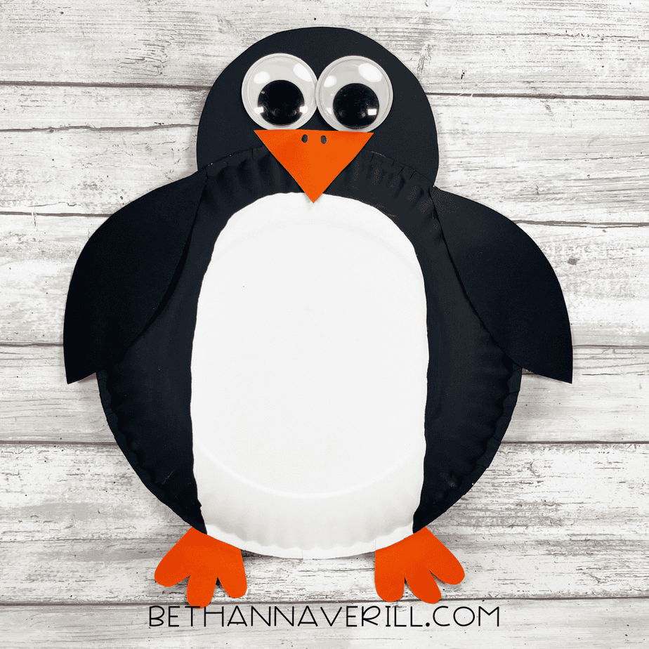 Completed paper plate penguin craft with black wings, big googly eyes, orange triangular beak, and orange feet on a wooden background.