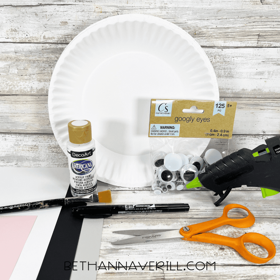 A flat lay of polar bear paper plate craft supplies including a white paper plate, googly eyes, white paint, black and pink cardstock, scissors, a glue gun, paintbrushes, and a black permanent marker on a rustic wooden background.