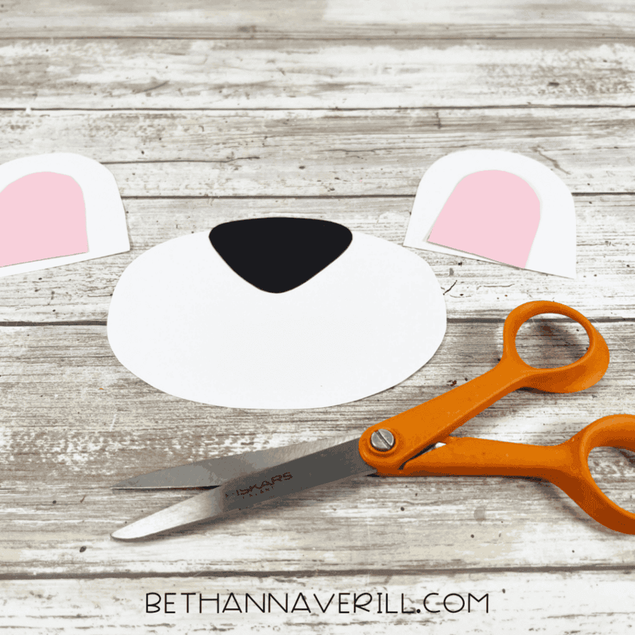 Cut-out cardstock pieces for the polar bear craft—white ears with pink centers and a large white muzzle with a black nose—next to orange scissors on a wooden surface.