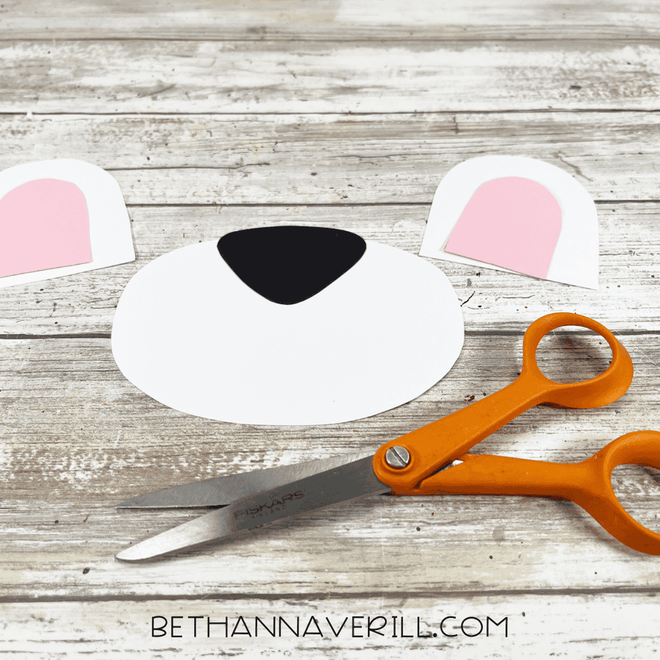 Cut-out cardstock pieces for the polar bear craft—white ears with pink centers and a large white muzzle with a black nose—next to orange scissors on a wooden surface.