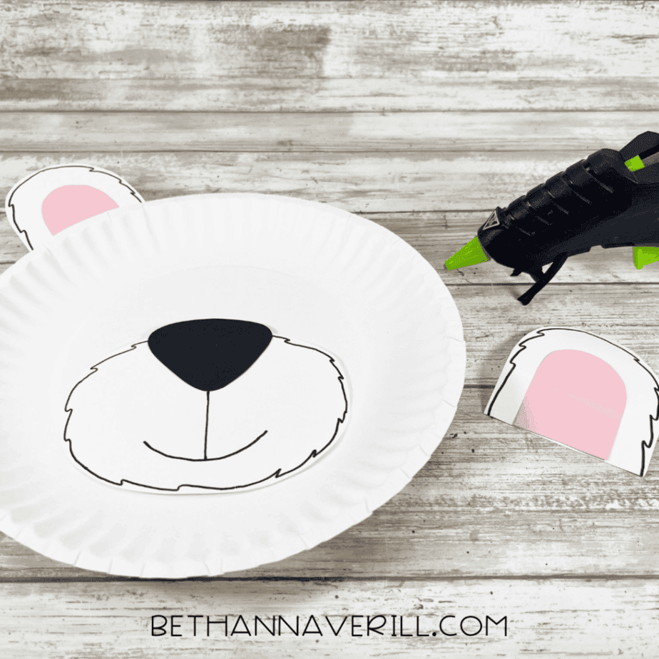 A white paper plate with the polar bear’s muzzle glued to the center and one ear already attached, with a glue gun laying beside the craft.