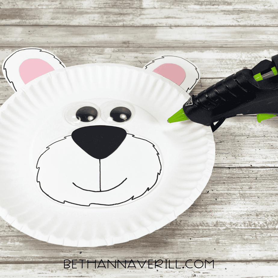 Googly eyes being glued above the polar bear’s muzzle on the paper plate, with the glue gun positioned nearby.