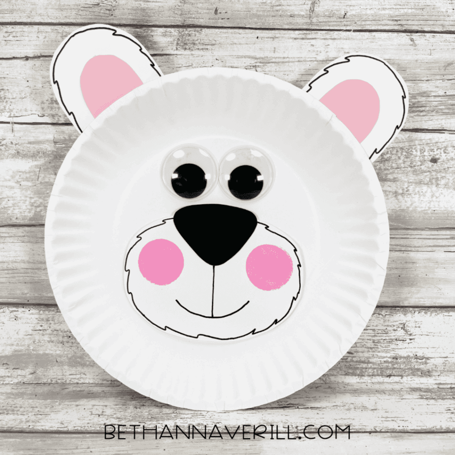 Completed polar bear paper plate craft featuring oversized googly eyes, pink cheeks, black line details, and cute rounded ears, displayed on a wooden background.