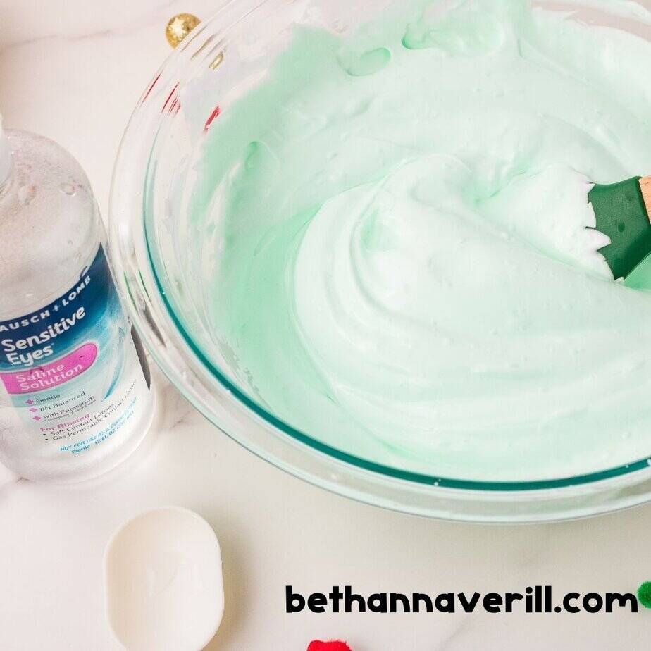 A bowl of smooth green slime sits next to a bottle of saline solution and a white measuring spoon, mid-process in making Grinch slime.