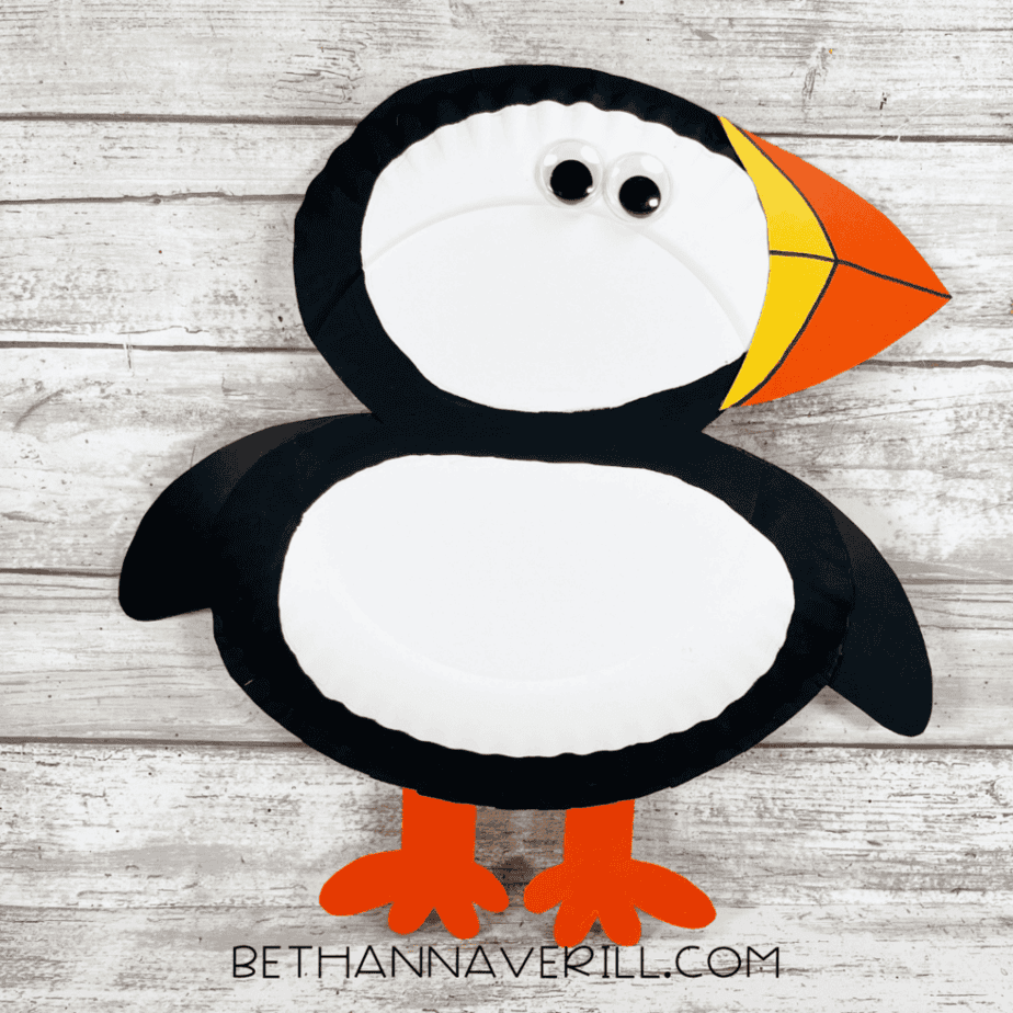 The completed puffin paper plate craft with a white belly, black wings and outline, bright orange feet, and a multicolored puffin beak.