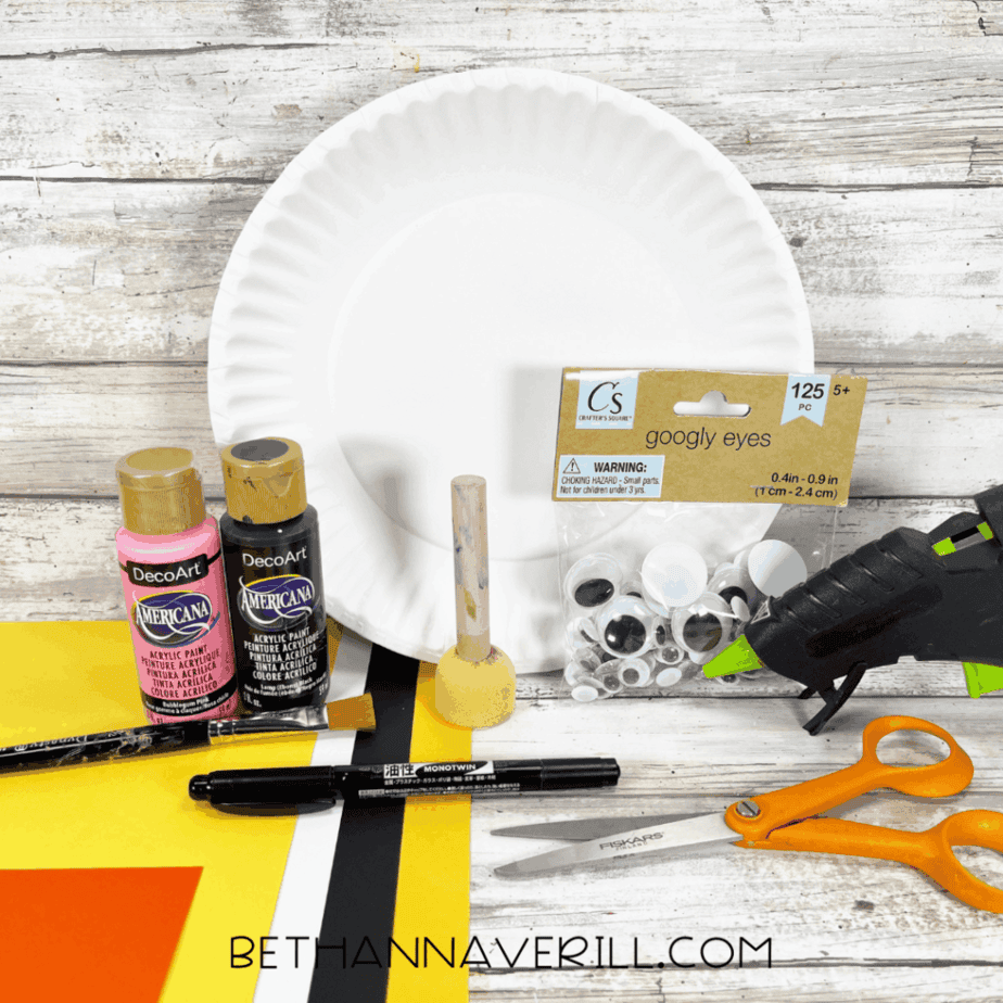 A white paper plate sits on a wooden surface alongside pink, black, and yellow acrylic paints, googly eyes, a hot glue gun, scissors, a black marker, a pencil, a paintbrush, a foam dauber, and sheets of orange, yellow, white, and black cardstock.