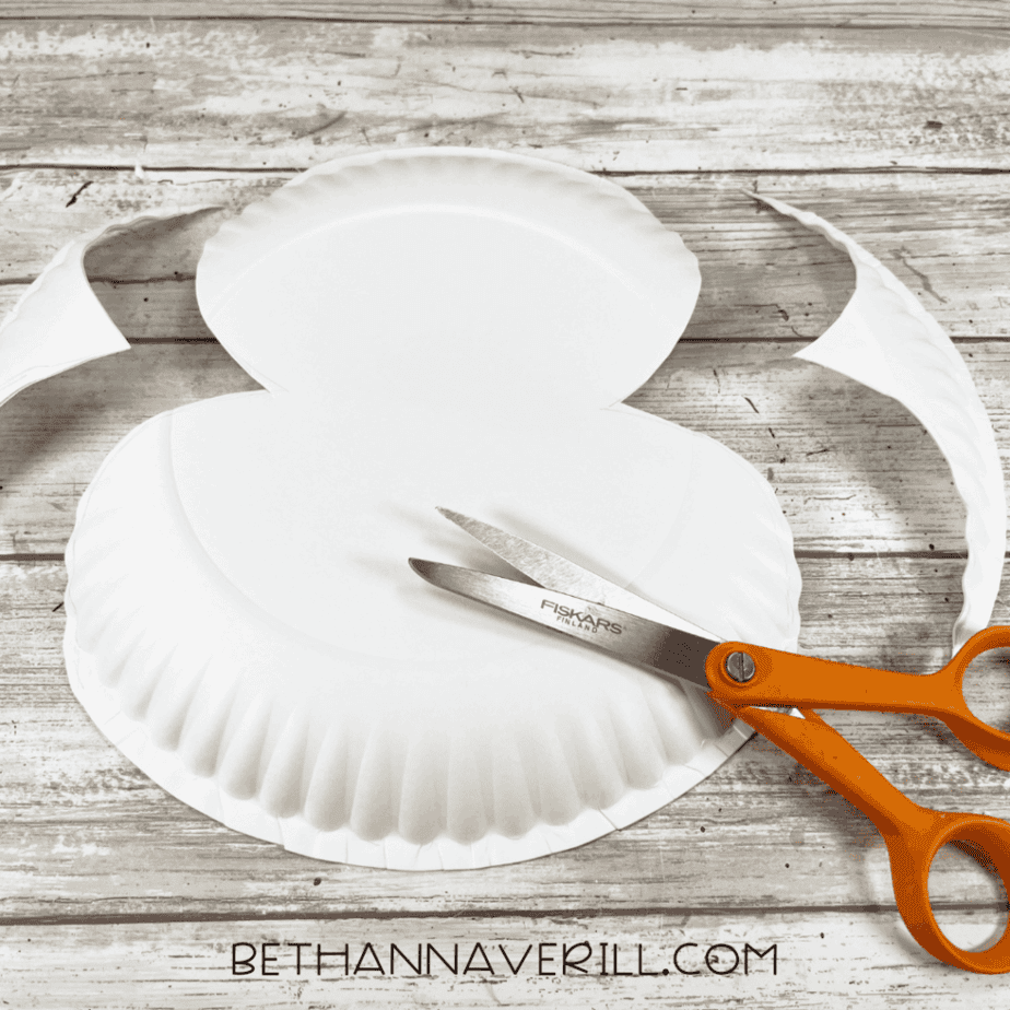 A paper plate cut into a two-circle puffin body shape, with the trimmed rim pieces pushed aside and scissors resting next to it.
