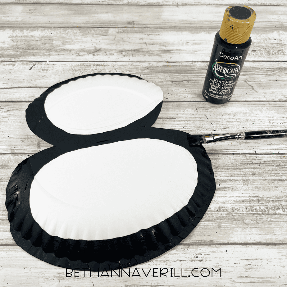 The cut paper-plate puffin shape partially painted black around the edges while the centers remain white. A black bottle of paint and paintbrush sit nearby.