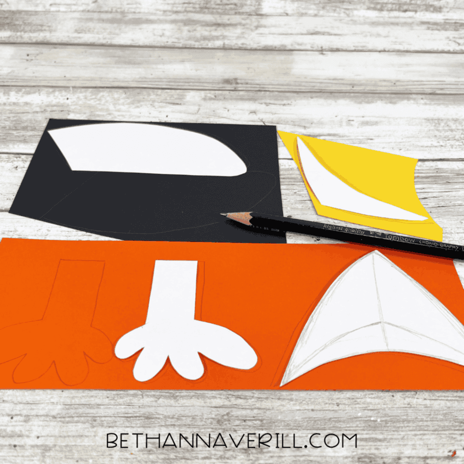 Cardstock pieces being traced with pencil using puffin beak, wings, and foot templates. Black, yellow, and orange paper are spread across the table.