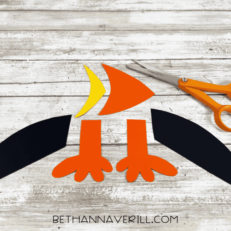 Cut-out puffin pieces — two black wings, two orange feet, and the layered orange-and-yellow beak — arranged on the table with scissors beside them.