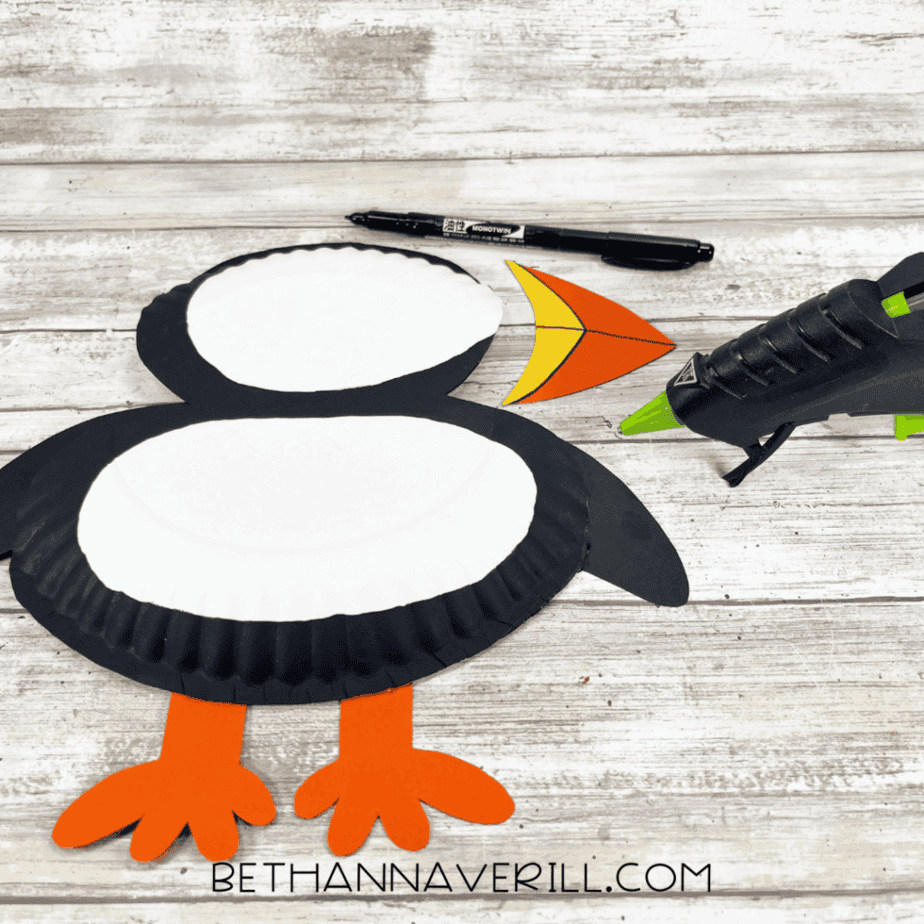 The puffin craft being assembled: wings and feet glued to the painted paper-plate body, with the beak and a glue gun placed near the top.