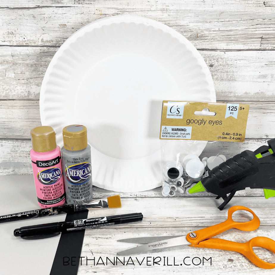 Craft supplies for a seal paper plate craft, including a white paper plate, googly eyes, gray and pink acrylic paint, scissors, glue gun, and cardstock arranged on a wooden background.