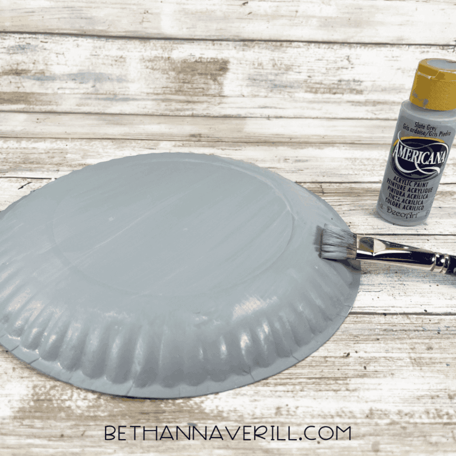 The back of a paper plate painted solid gray with a flat paintbrush and gray acrylic paint beside it.