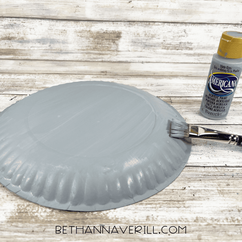 The back of a paper plate painted solid gray with a flat paintbrush and gray acrylic paint beside it.