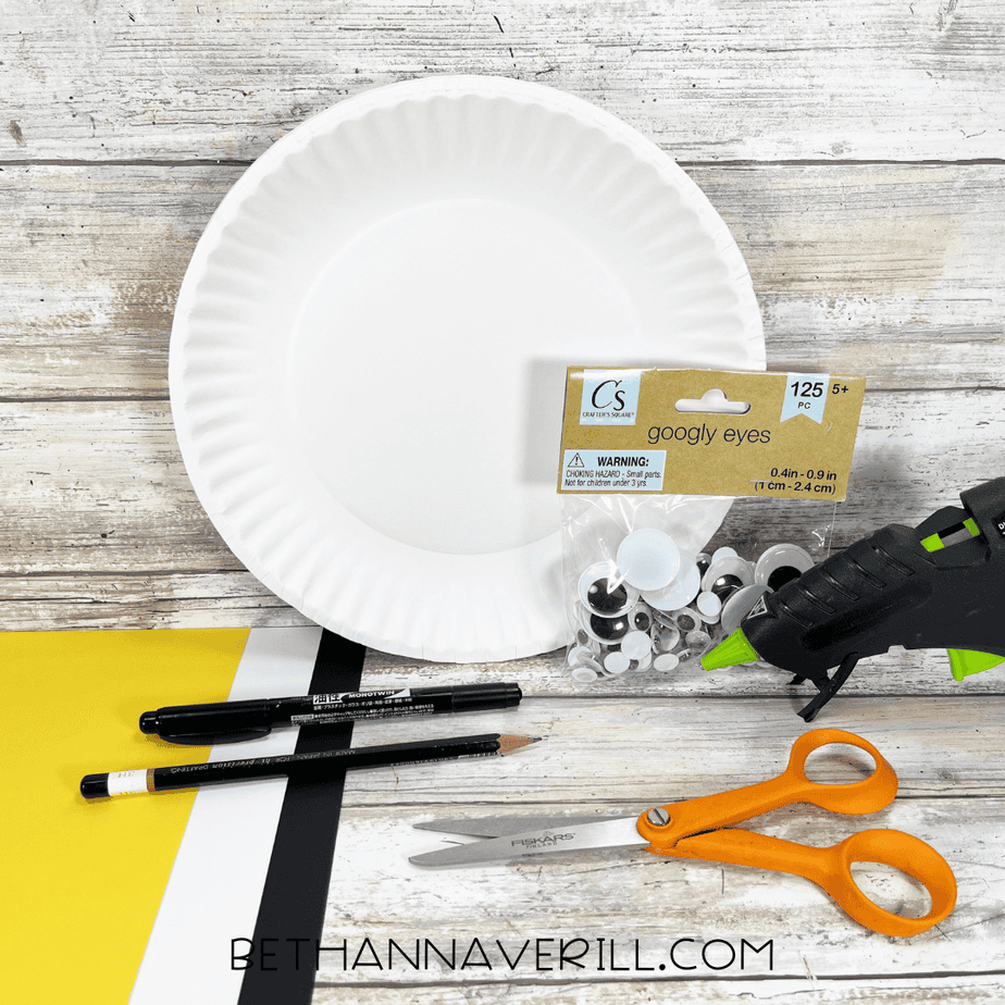 Paper plate snowy owl craft supplies including a white paper plate, googly eyes, yellow and black cardstock, pencil, markers, scissors, and hot glue gun on a wooden table.