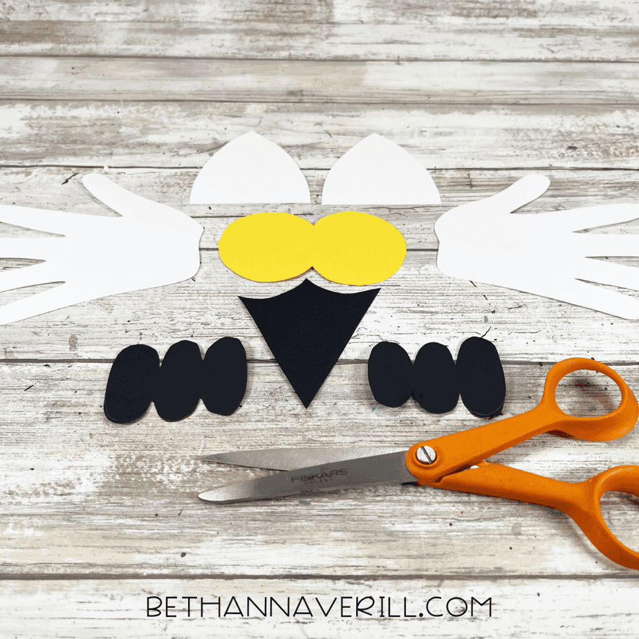 Cut-out snowy owl craft pieces including handprint wings, yellow eye circles, black beak, and black feet arranged on a wooden table with scissors.