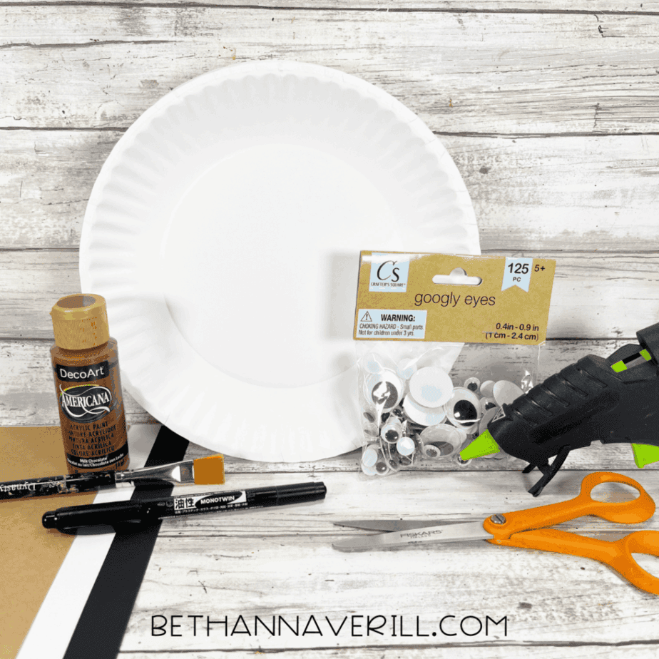 Supplies for a walrus paper plate craft, including a white paper plate, brown paint, googly eyes, scissors, glue gun, paintbrush, markers, and cardstock.