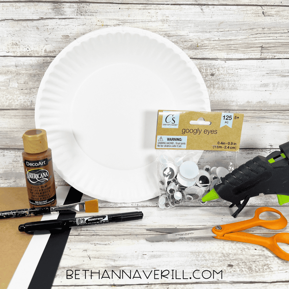 Supplies for a walrus paper plate craft, including a white paper plate, brown paint, googly eyes, scissors, glue gun, paintbrush, markers, and cardstock.